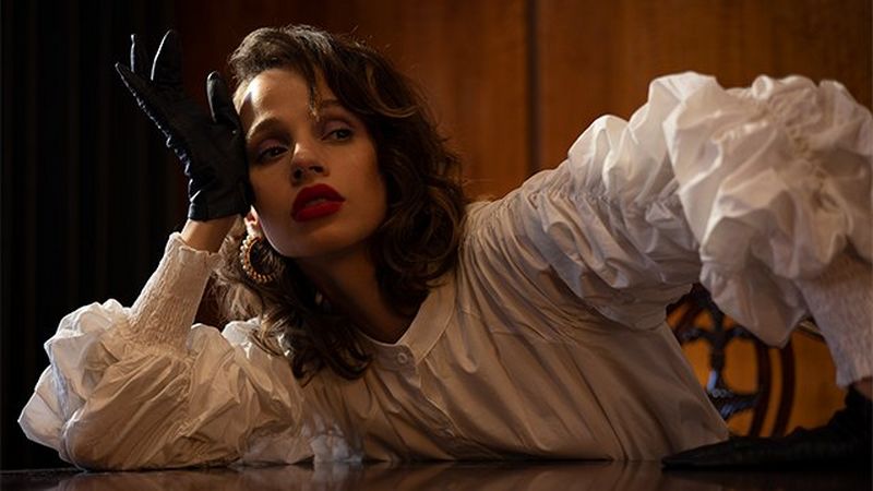 A woman in a white ruffled top and black gloves leans to her right on a desk in a darkened room, forming a triangular shape.