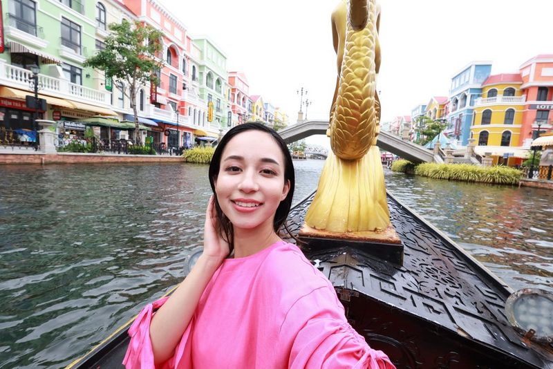 A person in a pink top holds their arm out to capture themselves travelling on a boat, with colourful buildings on either side, taken on a Canon EOS R50 with a Canon RF-S 10-18mm F4.5-6.3 IS STM lens.