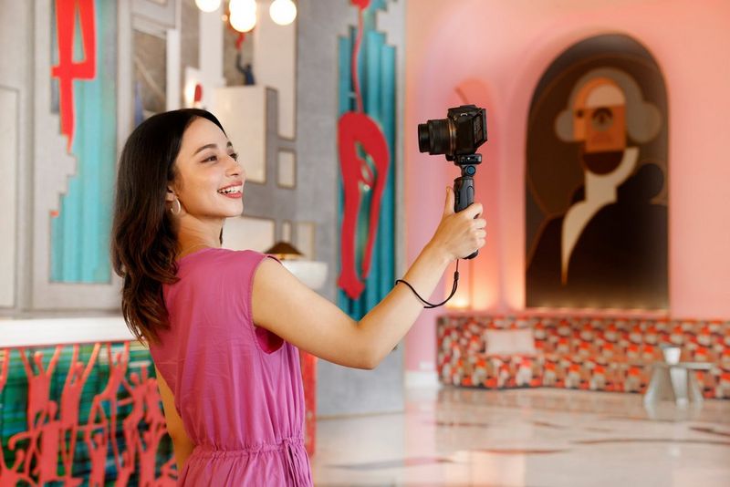 A woman in a pink dress holds a Canon camera with a Canon RF-S 10-18mm F4.5-6.3 IS STM lens in front of her using a tripod grip, smiling into the lens.
