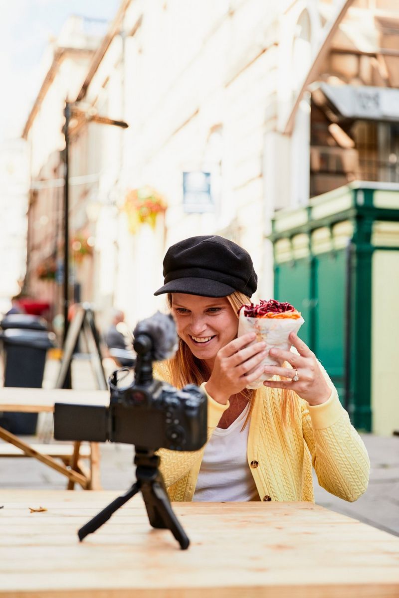 A young woman sitting at a table, smiling and holding a vegetable-stuffed wrap up to her camera, which sits on the table on a Canon Tripod Grip HG-100TBR in mini tripod form. 