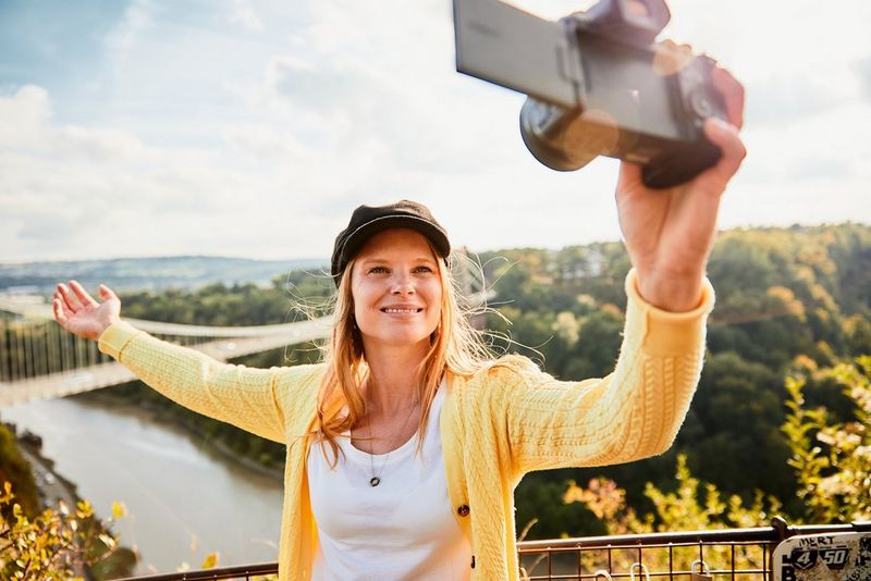 A young woman standing in front of a city skyline films herself on a Canon EOS R6 with a Canon RF 16mm F2.8 STM lens.