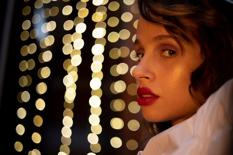 A low-light portrait of a woman in side profile, with blurred discs of light in the background, taken with a Canon RF 50mm F1.8 STM lens.