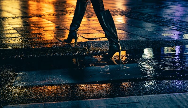 A person in high heeled boots walking through a puddle at night, orange and blue lights reflected on the wet pavement.