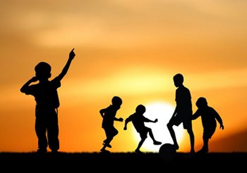 The silhouettes of four children, all different heights, play football against a dramatic orange sunset. The contrast between the flat blackness of the children and the deep orange of the sky makes them look like cardboard cut-outs.