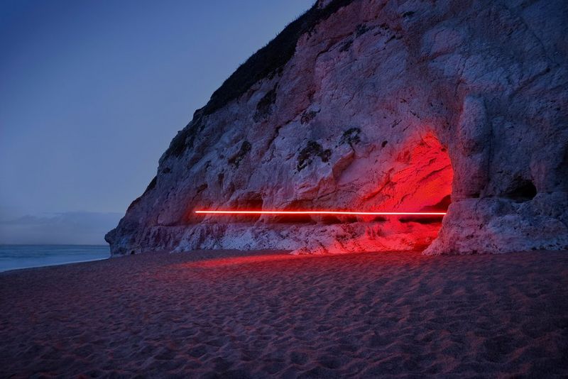 A long-exposure image of a horizontal red light trail emerging from a cave in a cliff face on a sandy beach.