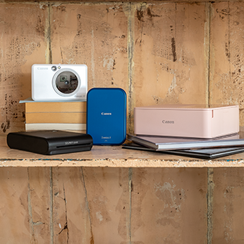 A collection of compact Canon photo printers in various colours on a wooden shelf. 