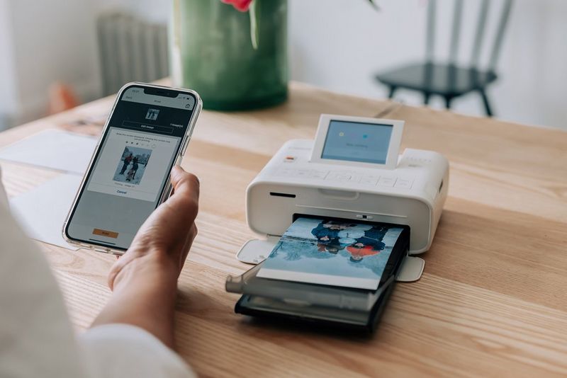 A person holding a phone with the picture of two people on it. SELPHY printer in the background with the same picture being printed.