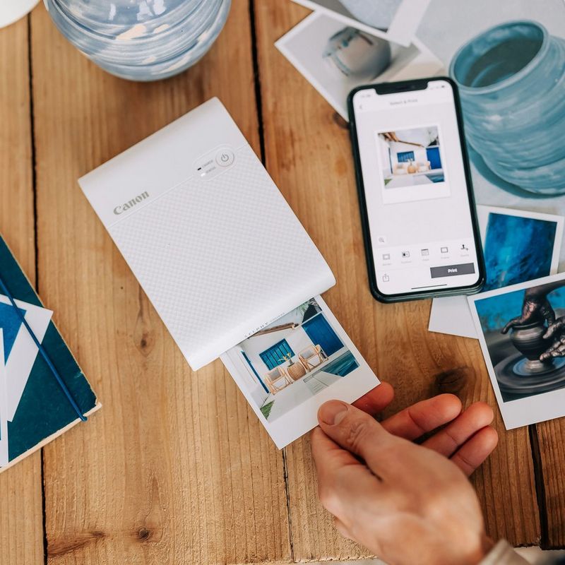 White SELPHY Square QX10 printing a picture of a white house with blue wooden window shutters. A hand is holding the edge of the photo. Phone is next to it showing the same picture. Some other printed pictures are scattered on the table.