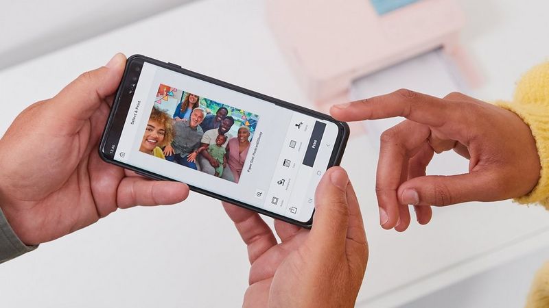 Two hands holding a phone with a picture of a family with stamps applied in SELPHY Photo Layout app. Another hand is visible in the top right corner pointing at the phone.