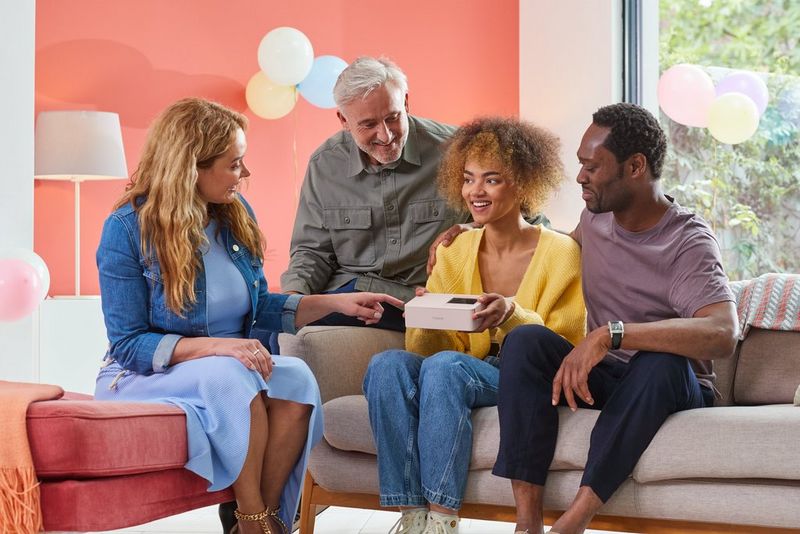 Four people sitting on sofas in a decorated room with balloons in the background. One of them, a young girl is holding SELPHY CP1500 in pink and others looking and pointing at the printer.
