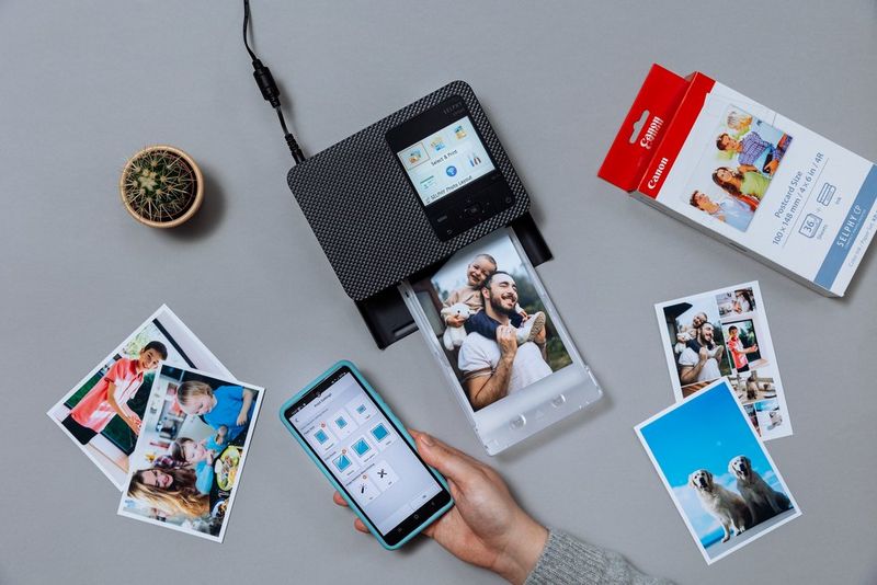 A Canon SELPHY CP1500 printer with a family photograph on the print tray. Next to it are a selection of photo prints, a pack of printer paper and a hand holding a smartphone, displaying the SELPHY Photo Layout app.