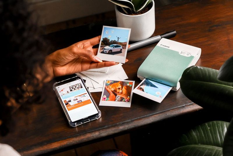 A pair of hands holds a smartphone showing a photo with white border on the screen, while a Canon SELPHY Square QX10 sits on a table in the background. 