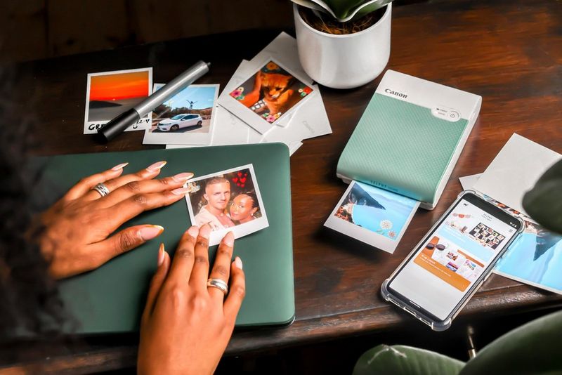 A Canon SELPHY Square QX10 sits on a table printing out a holiday photo. Next to it a pair of hands smooth a sticker onto a laptop, and to the other side a smartphone shows the SELPHY Print Layout app.