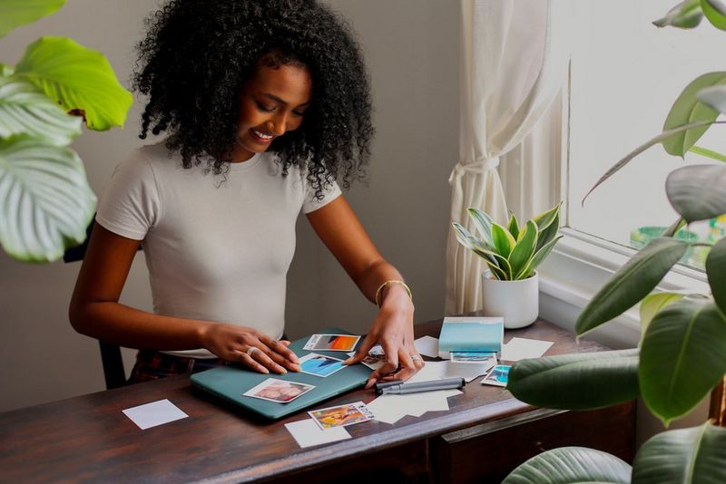 A woman with black curly hair smiles as she sticks photos printed on a Canon SELPHY Square QX10 onto the lid of her laptop.