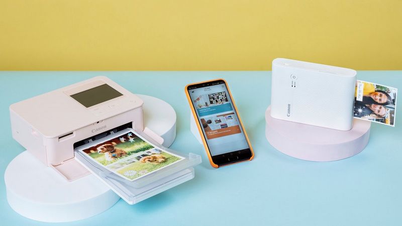 White SELPHY Square QX10 printer and pink SELPHY CP1500 printer and a mobile phone with SELPHY Photo Layout app between them.