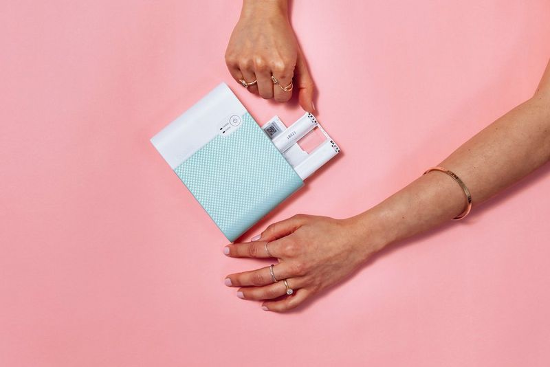 A pair of hands slot in the ink cartridges to the side of a Canon SELPHY Square QX10 printer.