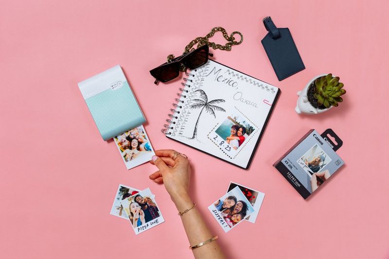 A Canon SELPHY Square QX10 printer sits on a pink background, surrounded by prints, a scrapbook and accessories. A hand is removing a print from the printer.