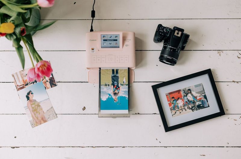 A pale pink Canon SELPHY CP1300 printer on a white wooden surface, printing an image, surrounded by family photos, some with frames and some without, a Canon EOS 250D camera and some pink flowers.