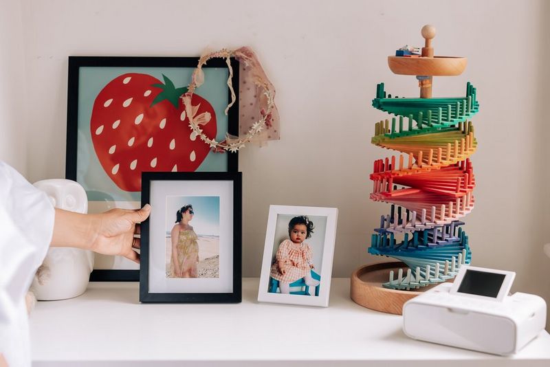 Two framed portrait photos on a low shelf beside a picture of a strawberry, a rainbow-coloured toy and a Canon SELPHY CP1300.
