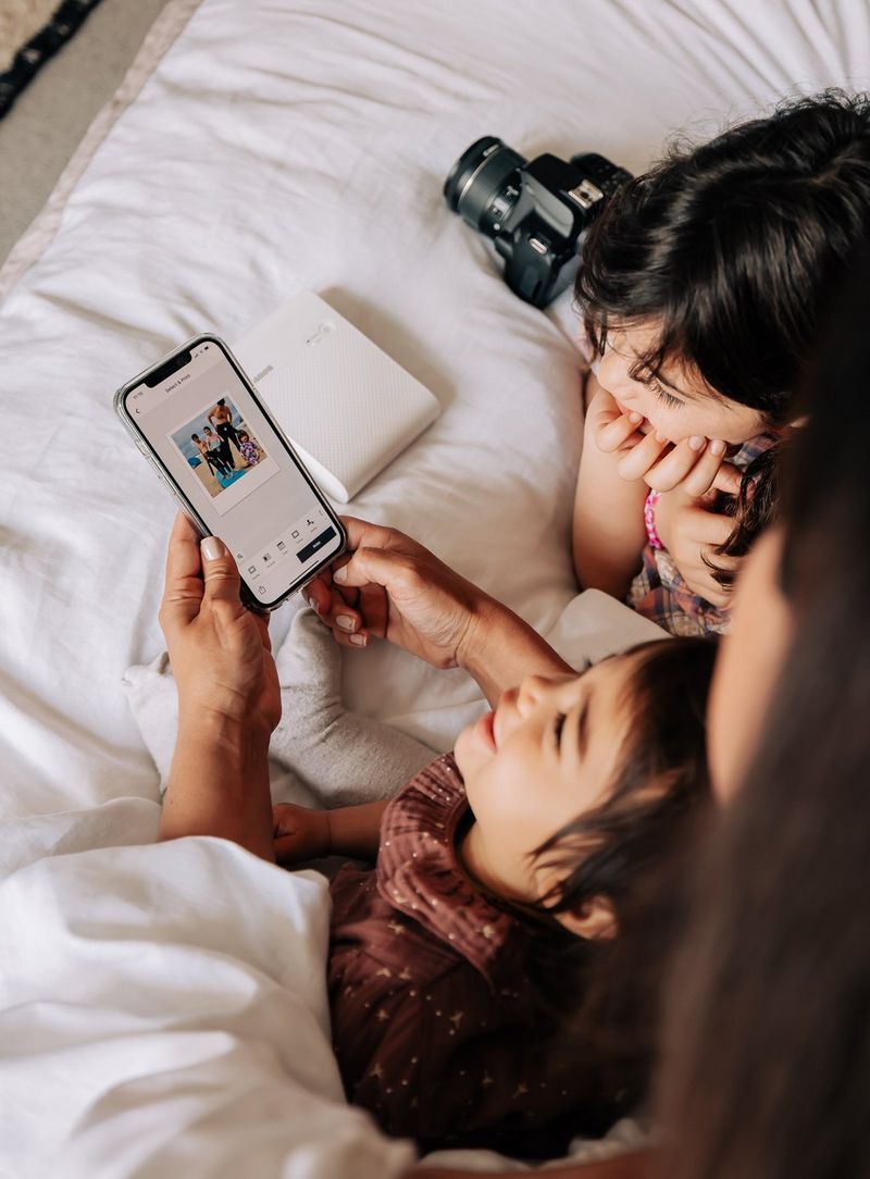 Two young children sitting on a bed with a Canon SELPHY Square QX10, a Canon EOS 250D and their mother, who is holding a phone with the SELPHY Photo Layout app open.