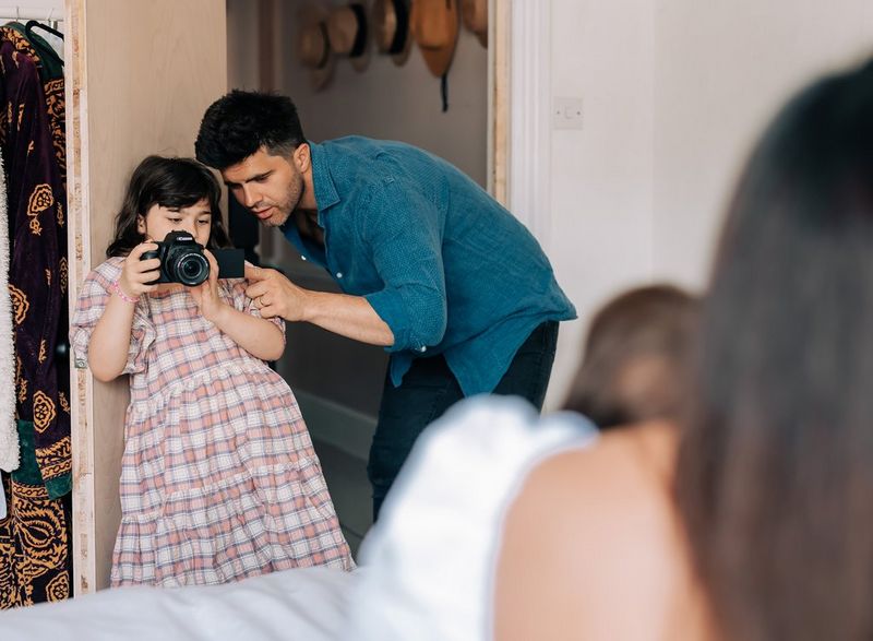 A man helping his daughter photograph the rest of their family using a Canon EOS 250D camera.