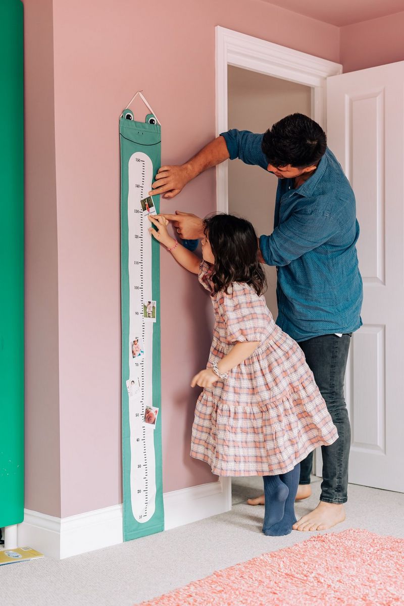 A man helping his daughter stick a photograph onto a height chart shaped like an elongated frog.