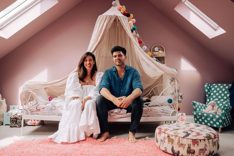 A couple smile at the camera while sitting on a child's bed in an attic bedroom.