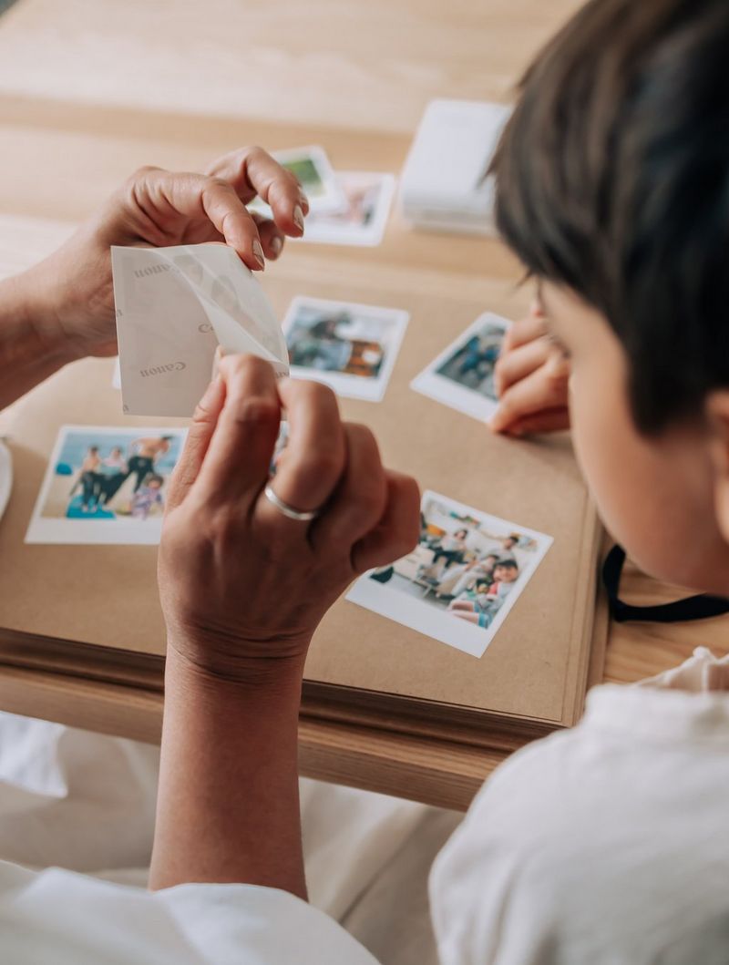 A woman's hand peeling off the backing from a printed photo to reveal the sticky side beneath, preparing to stick it to the cover of a scrapbook.