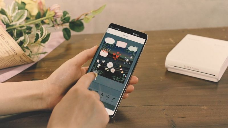 A person holding a phone with stamps available on SELPHY Photo Layout app showing on it. One hand is tapping on the screen. In the background we can see white SELPHY Square QX10 on a table.