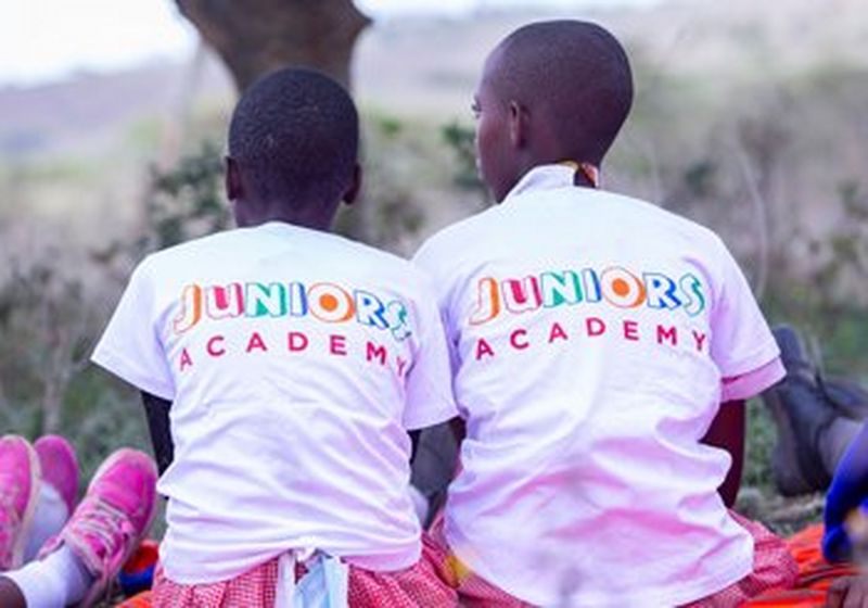 Two children, photographed from behind. They are sat side-by-side and wear white t-shirts with ‘Juniors Academy’ written across the back in bright, primary colours and red and white checked skirts. Others sit beside them, but only their feet can be seen.