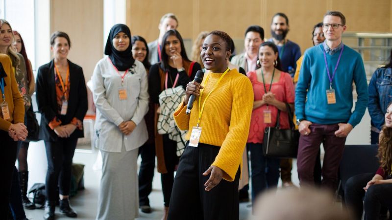 A woman in a yellow textured jumper uses a microphone to talk to a mixed group of people in a lecture theatre. They are all wearing lanyards and standing in a wide circle around her. 