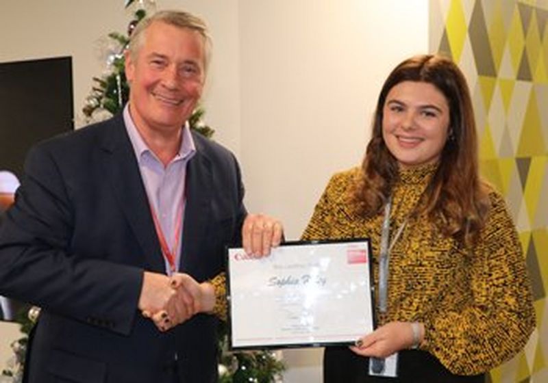 A smiling man and woman stand facing the camera, shaking hands. They are holding a framed certificate to the camera with their spare hands. The man is wearing a dark blue suit, and shirt with no tie. The woman is wearing a high-necked yellow and black blouse. They both wear lanyards around their necks.