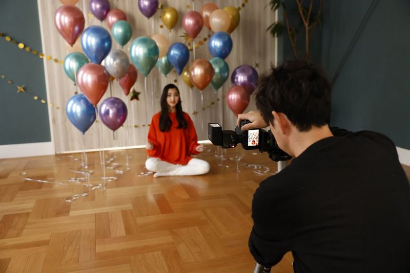 A man taking a picture with a Canon camera and Speedlite EL-10 flash of a woman sitting on the floor surrounded by balloons. He has the camera tilted for a vertical shot.