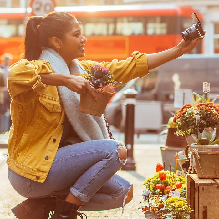 A woman crouches to street level to take a selfie with a plant, using a Canon camera.
