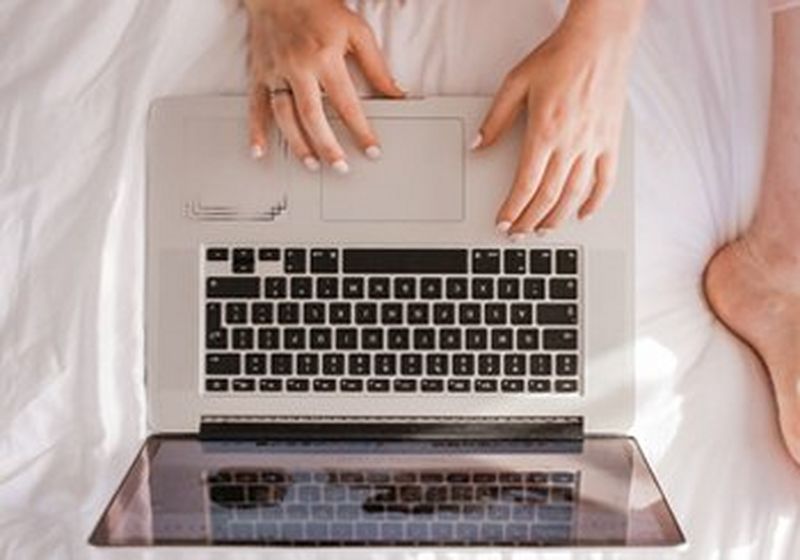 Laptop from above, on white bedsheets, with a woman’s hands and bare foot in shot