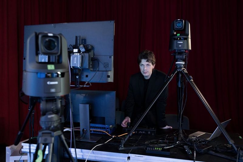 A man stands at a desk covered in electrical equipment, looking at two large monitors and with one hand on a controller in front of him. Two Canon PTZ cameras are on tripods, facing forwards from him.