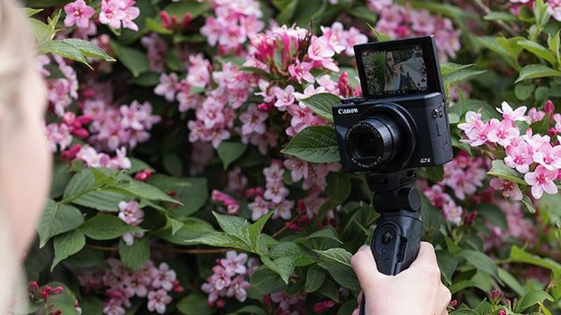 A blonde woman holding Canon PowerShot G7 X Mark III on a tripod in front of her, speaking to camera.