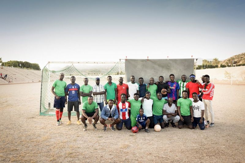 The grey photographer’s backdrop is pinned on the right half of a set of goalposts. Half in front of the goalposts and half in front of the backdrop are twenty young Black men posing in two rows, like a football squad, with the front row crouching. The ground beneath them is parched earth and the walls of a storm drain rise to the left and right. 