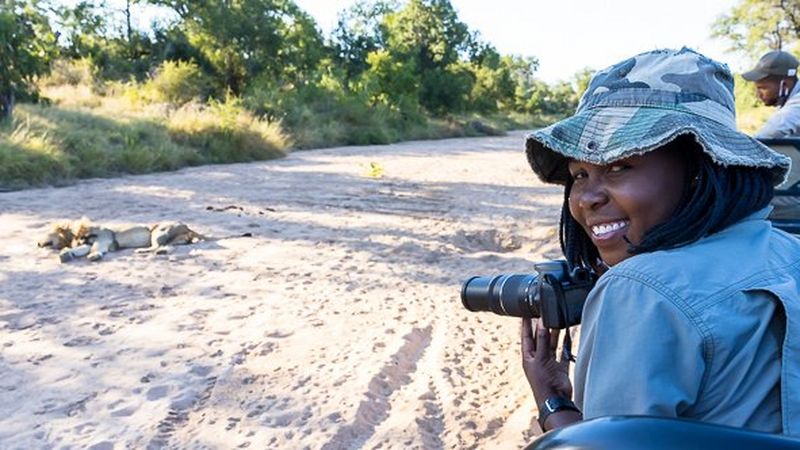 A young woman in a green camouflage hat and blue shirt turns her head to smile for the camera. She is holding a camera in her hand and it is pointed at a lion, which lays on the dirt road in front of her.