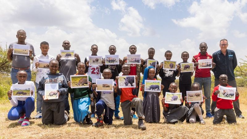 Twenty people line up in two rows, with the front row sitting or kneeling. They are on grass with a blue cloud-filled sky behind them. Each holds a printed picture of a rhino. 