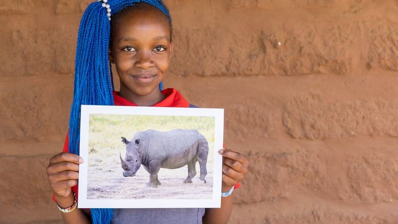 A child with long bright blue braids and a red t-shirt holds up a photograph of a rhino. She stands against a terracotta-coloured wall. 