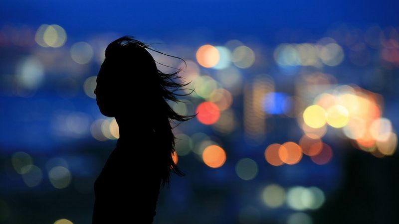 A silhouette of a woman with her hair flowing in the breeze, in front of bokeh city lights.