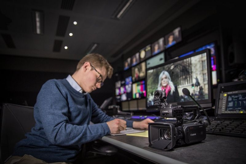 A journalist sits in front of a bank of TV screens. A Canon XF405 professional camcorder is on the desk beside him.