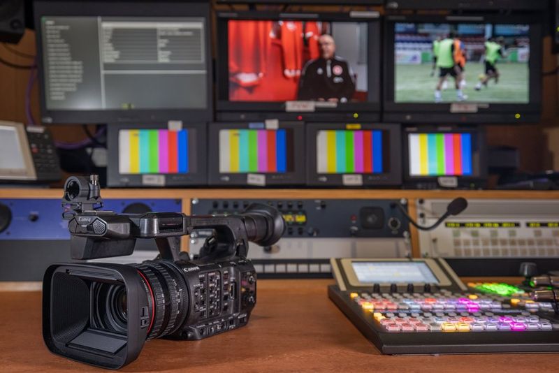 A Canon XF605 camcorder on a recording desk with rows of screens and panels behind it.