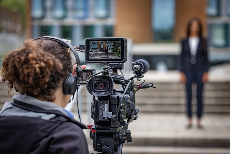 A person shot from behind filming a woman dressed in a suit and standing in front of a large building.
