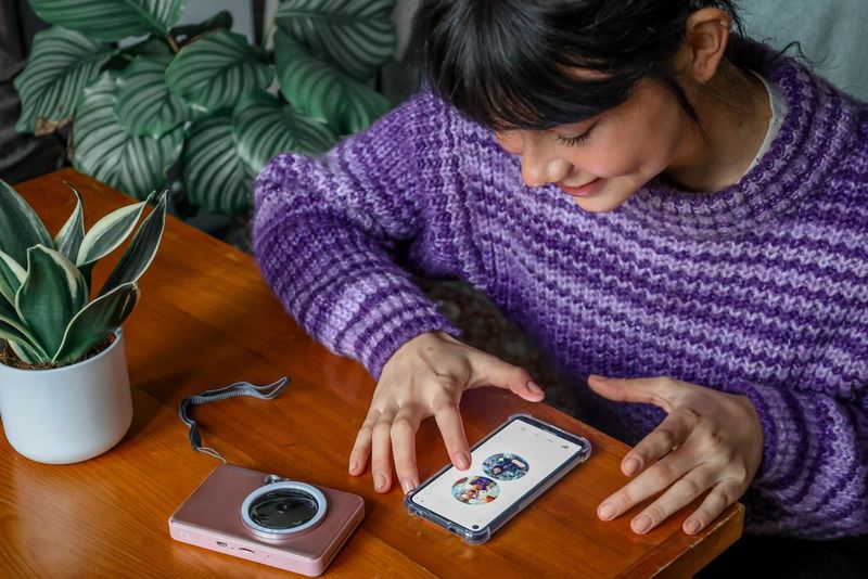 A woman in a purple jumper uses a Canon printing app to customise her photos while a Canon Zoemini S2 sits next to the smartphone on a table.