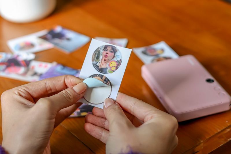 A pair of hands hold up a small sheet of two circular stickers, peeling one off. Behind them a Canon Zoemini S2 sits on a table surrounded by other photo prints.