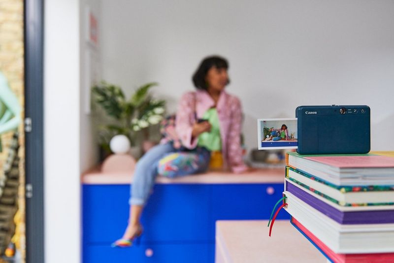 A woman sat on a blue cabinet, her Canon Zoemini S2 balanced on a pile of books in the foreground.