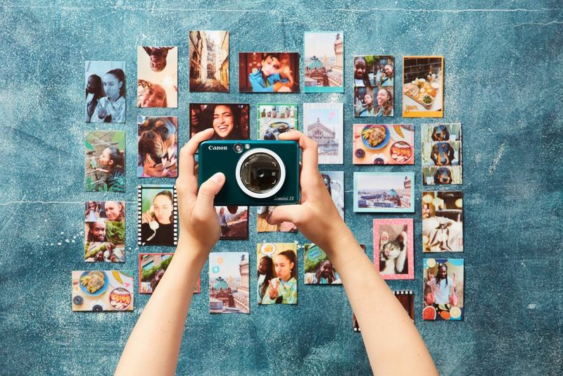 A person taking a photograph on a Canon Zoemini S2 of a wall containing lots of smaller photographs of two young people having fun.