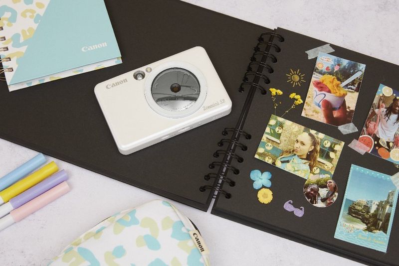 A desk with a scrapbook with black pages, a light-blue notebook and a white Canon Zoemini S2 photo printer on it. 